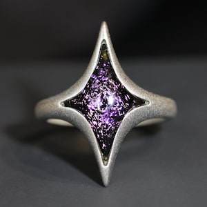 May include: A silver ring featuring a four-pointed star design. The star is filled with a purple, sparkly glass.