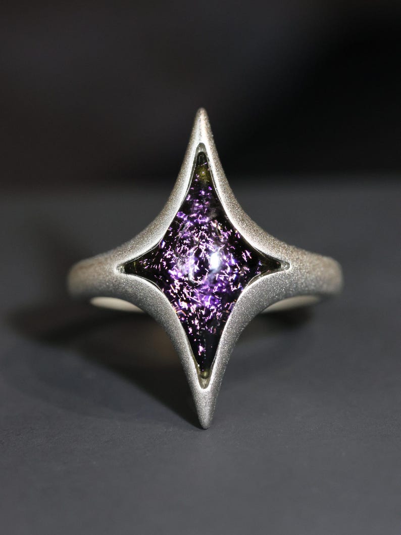 May include: A silver ring featuring a four-pointed star design. The star is filled with a purple, sparkly glass.