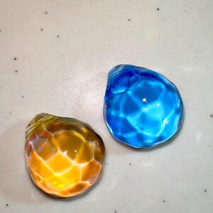 Puede incluir: Dos canicas de vidrio, una azul y otra naranja, con una superficie texturizada. Las canicas est&aacute;n sobre un fondo blanco.