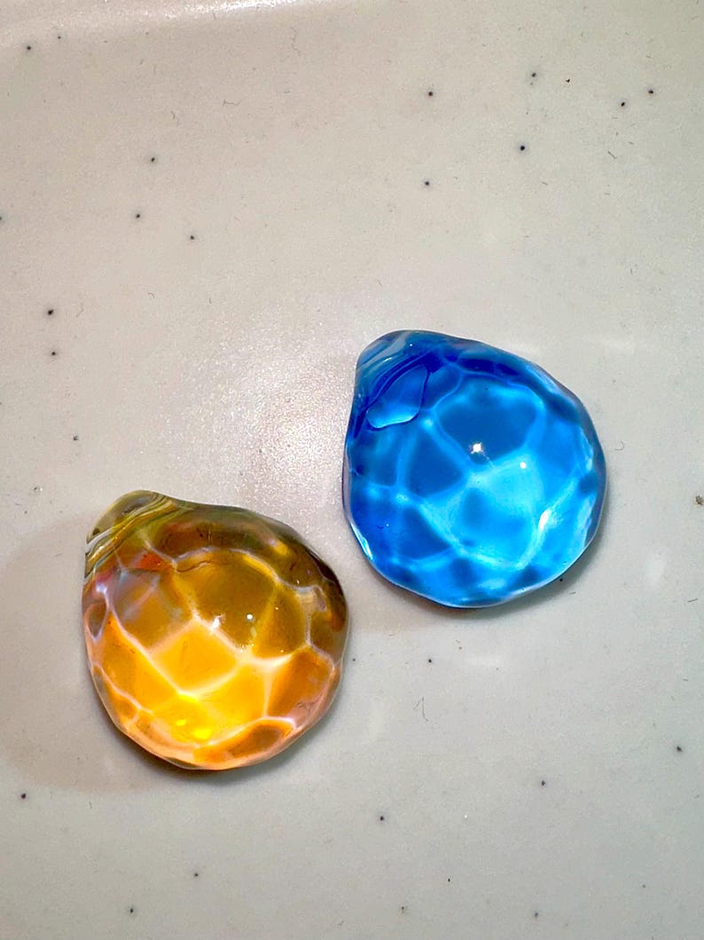 Puede incluir: Dos canicas de vidrio, una azul y otra naranja, con una superficie texturizada. Las canicas est&aacute;n sobre un fondo blanco.
