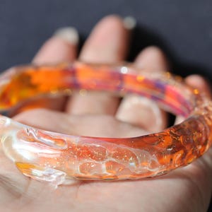 May include: A glass bangle bracelet with a swirling orange, yellow, and white pattern. The bracelet is made of clear glass with a textured surface.
