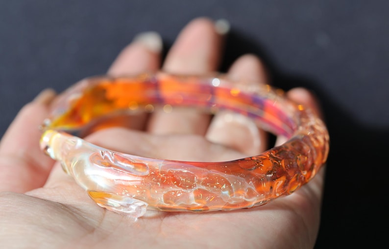 May include: A glass bangle bracelet with a swirling orange, yellow, and white pattern. The bracelet is made of clear glass with a textured surface.