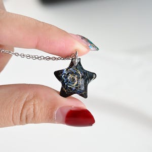May include: A silver chain necklace with a black star-shaped pendant. The pendant has a swirling, galaxy-like design with gold accents.