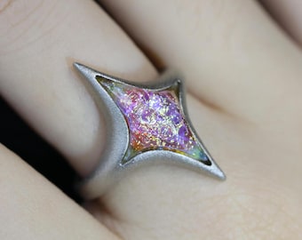 May include: A silver ring with a four-pointed star shaped setting. The star is filled with iridescent pink, purple, and gold glitter.