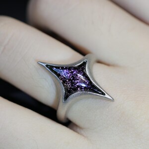 May include: A silver ring featuring a four-pointed star design. The star is filled with a dark purple, glittery material.