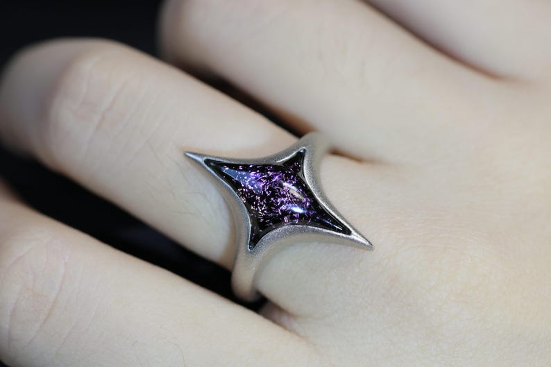 May include: A silver ring featuring a four-pointed star design. The star is filled with a dark purple, glittery material.