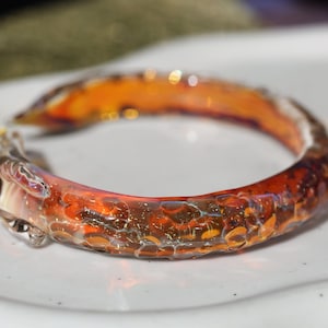 May include: A close-up of a glass bangle bracelet with a textured, orange and brown design. The bracelet is made of glass and has a unique, handmade look.