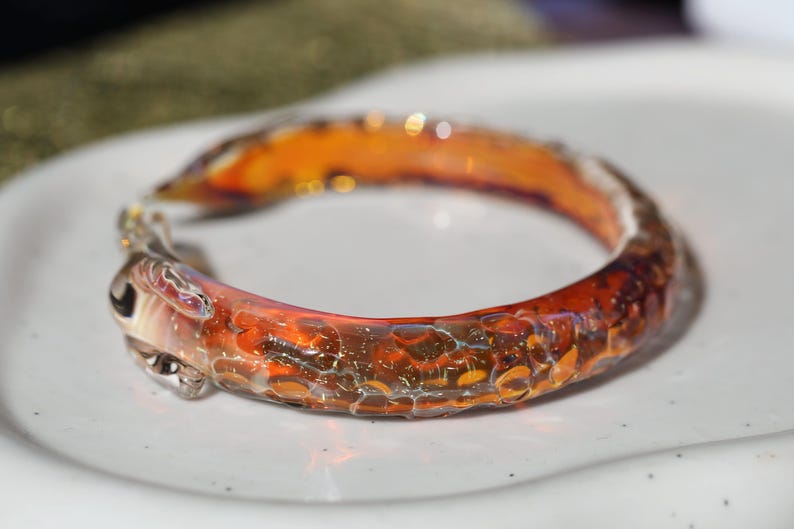 May include: A close-up of a glass bangle bracelet with a textured, orange and brown design. The bracelet is made of glass and has a unique, handmade look.