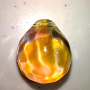 Puede incluir: Un colgante de vidrio amarillo con un patr&oacute;n de remolino iridiscente. El colgante es liso y redondeado, con una parte superior ligeramente puntiaguda.