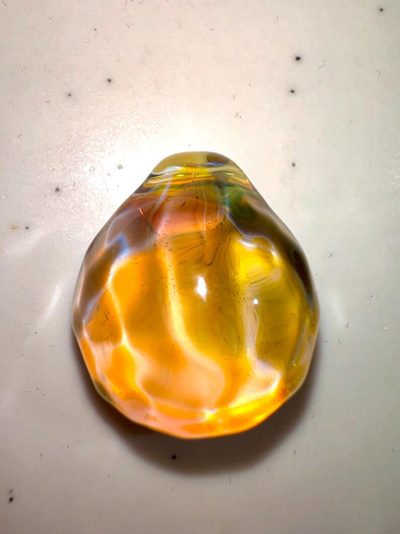 Puede incluir: Un colgante de vidrio amarillo con un patr&oacute;n de remolino iridiscente. El colgante es liso y redondeado, con una parte superior ligeramente puntiaguda.