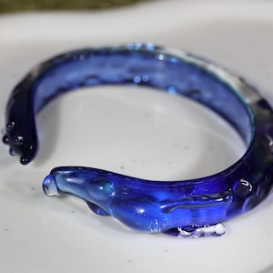 May include: A blue glass bangle bracelet with a textured, wavy design. The bracelet is made of clear glass with a blue tint.