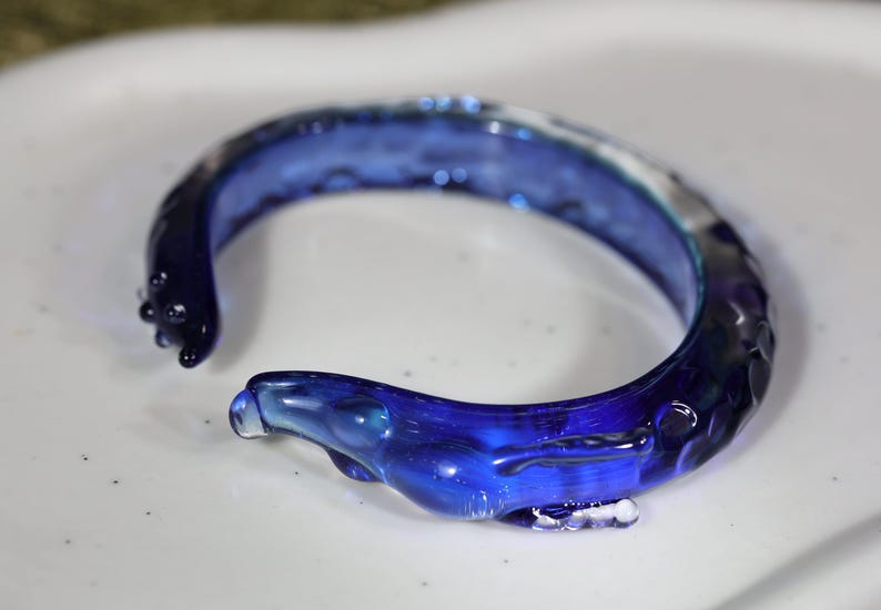 May include: A blue glass bangle bracelet with a textured, wavy design. The bracelet is made of clear glass with a blue tint.