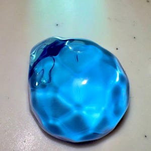 Puede incluir: Una gema de vidrio azul brillante, facetada, con una forma ligeramente irregular. La gema es transl&uacute;cida y refleja la luz.