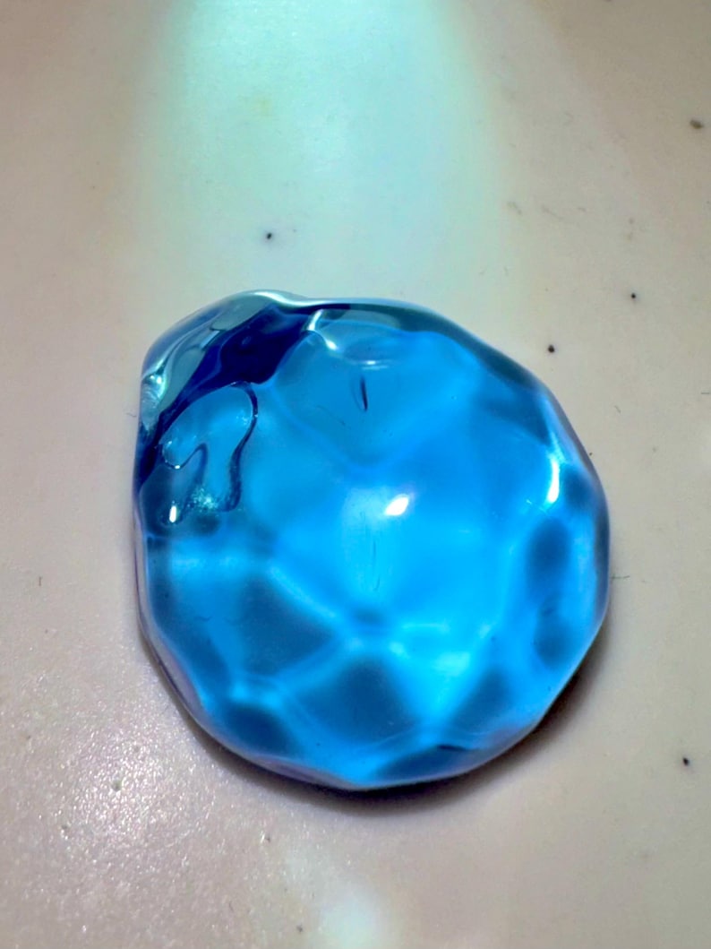 Puede incluir: Una gema de vidrio azul brillante, facetada, con una forma ligeramente irregular. La gema es transl&uacute;cida y refleja la luz.