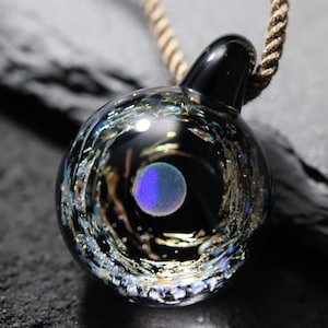 Pode incluir: Um colar com um pingente de vidro preto que representa uma galáxia em espiral com um planeta azul e roxo no centro.