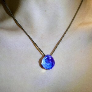 Puede incluir: Un collar con un colgante de vidrio azul en un cord&oacute;n marr&oacute;n oscuro. El colgante es redondo, de forma irregular, con un patr&oacute;n de remolino y nublado.