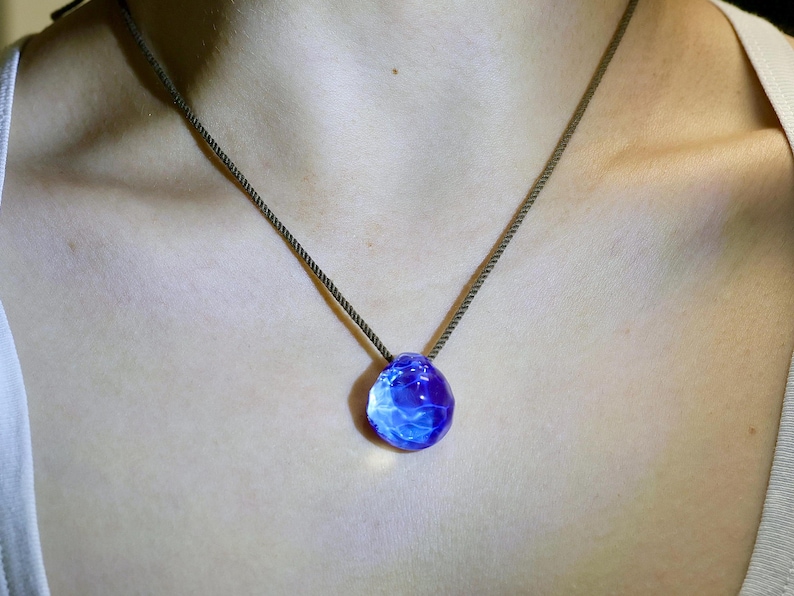 Puede incluir: Un collar con un colgante de vidrio azul en un cord&oacute;n marr&oacute;n oscuro. El colgante es redondo, de forma irregular, con un patr&oacute;n de remolino y nublado.