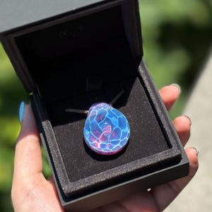 Könnte beinhalten: Eine tropfenförmige Glaskette mit einem blauen und rosa schillernden Finish. Der Anhänger hat eine strukturierte Oberfläche und wird in einer schwarzen Schachtel präsentiert.