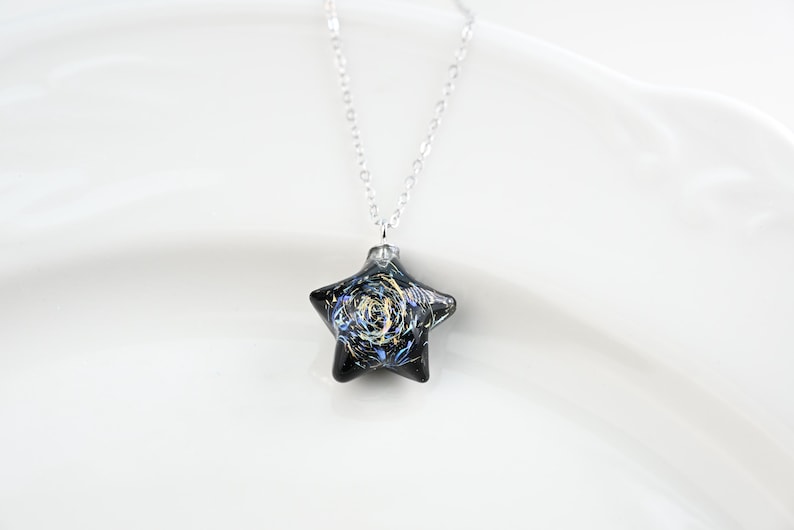 May include: A silver chain necklace with a black star-shaped pendant. The star has a swirling pattern of gold and blue paint.