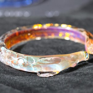 May include: A glass bangle bracelet with a iridescent finish. The bracelet has a unique, organic shape and is made of clear glass with hints of orange, purple, and white.