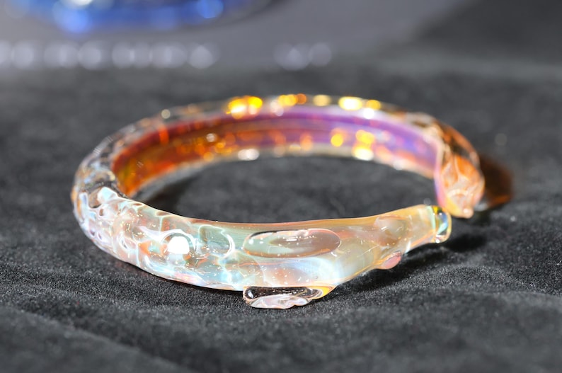 May include: A glass bangle bracelet with a iridescent finish. The bracelet has a unique, organic shape and is made of clear glass with hints of orange, purple, and white.