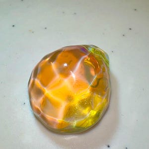 Puede incluir: Una esfera de vidrio transparente y facetada con un brillo iridiscente amarillo y naranja.