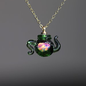 May include: A gold chain necklace featuring a green glass teapot pendant. The teapot pendant has a colourful, iridescent stone inside. The teapot has a handle and a spout.