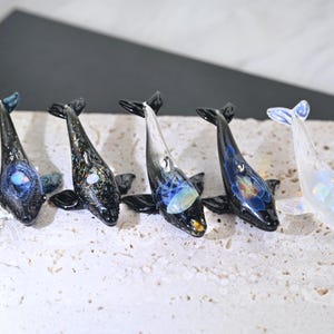 May include: Five glass orca figurines with black and iridescent bodies, and blue or white fins and tails. Each whale has a unique design, with some featuring internal patterns of colour. The figurines are displayed on a textured, light-coloured surface.