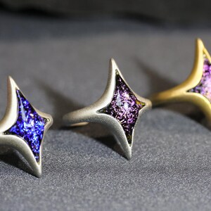 May include: Three silver rings with a star-shaped design. Each ring features a different colour of iridescent glass in the centre of the star: blue, purple, and pink.