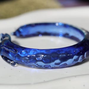 May include: A blue glass bangle with a textured surface. The bangle is made of clear glass with a blue tint and has a bumpy, uneven texture.