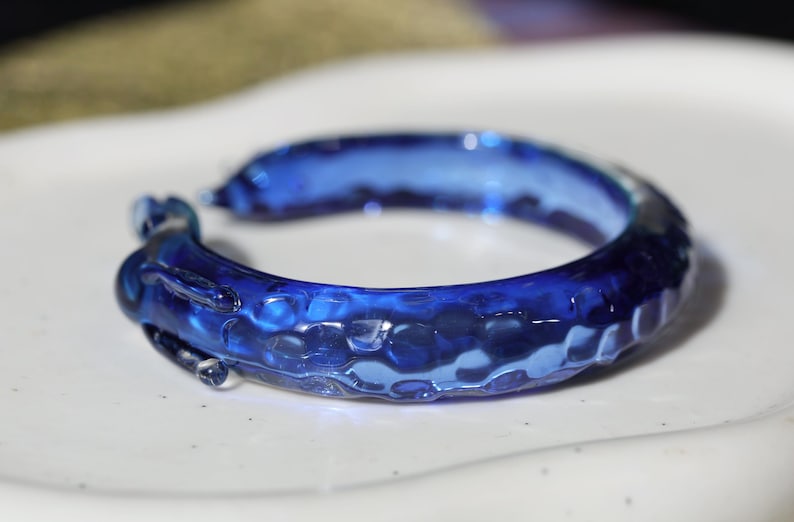 May include: A blue glass bangle with a textured surface. The bangle is made of clear glass with a blue tint and has a bumpy, uneven texture.
