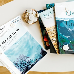 Pode incluir: Uma pilha de livros infantis sobre o oceano, incluindo "Over and Under the Waves" e "Sea Glass". Há também uma folha de papel com o título "Ocean Unit Study" e uma pequena cesta de conchas brancas.