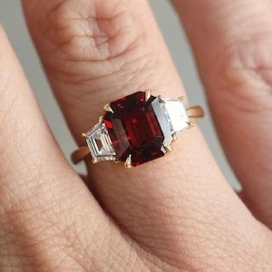 Puede incluir: Un anillo de oro con una gran piedra preciosa roja de talla esmeralda rodeada de dos piedras más pequeñas, transparentes, de talla baguette.