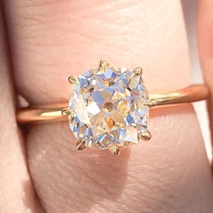 Puede incluir: Un anillo de oro con una gran piedra preciosa de talla cojín, transparente. El anillo está en un dedo.