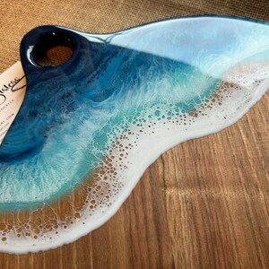 Puede incluir: Una tabla de servir de madera con un diseño de ola de océano de resina azul, verde y blanca. La tabla tiene un asa con un agujero. El texto "Salty Sisters" es visible en una tarjeta de visita en la imagen.