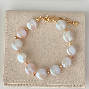 May include: A gold-toned bracelet featuring large, irregularly shaped white pearls with a subtle pink hue. Small gold beads separate each pearl. The bracelet is on a light beige surface.