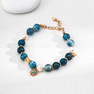 May include: A beaded bracelet featuring blue and white round beads, interspersed with white pearls and gold-colored accents. A small gold charm with a turquoise center adds detail. The bracelet is displayed on a white surface.
