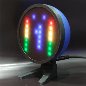 Puede incluir: Una pantalla LED redonda, azul y negra con un número azul "1" y dos filas verticales de luces de colores. La pantalla está en un soporte negro.