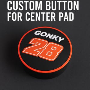Op de afbeelding: Een zwarte aangepaste knop voor een middenpad met de tekst "CUSTOM BUTTON FOR CENTER PAD" in het wit. De knop heeft een zwarte voorkant met het woord "GONKY" in het wit en het nummer "28" in het oranje met een witte rand. De knop heeft een oranje rand.