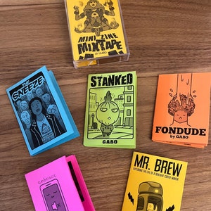 以下が含まれることがあります： イラストとテキストが描かれたカラフルな表紙のミニジンが5冊セットになっています。ジンは「The Sneeze」、「Stanked」、「Fondude」、「Embrace Silence」、「Mr. Brew」というタイトルです。