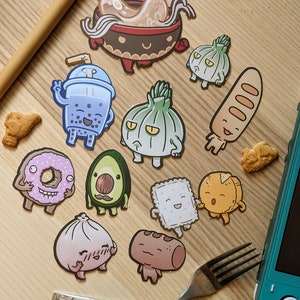 以下が含まれることがあります： ドーナツ、アボカド、ラーメン、バゲットなど、さまざまな食べ物を描いた、カラフルな漫画の食べ物ステッカー12個セット。ステッカーは、フォークとスプーンのある木製の表面に置かれています。