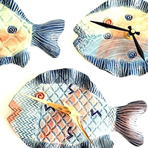 May include: Three fish-shaped wall clocks featuring hand-painted ceramic bodies with blue, peach, and white scales. Two clocks have black hands, while the third clock has gold hands. Each clock displays a unique pattern of swirls and geometric lines.