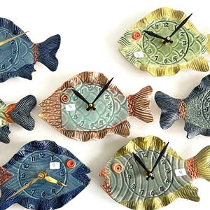 May include: A collection of seven ceramic fish-shaped wall clocks. Each clock features a different color scheme, including blue, green, and yellow, with gold-colored clock hands and numbers. The clocks are arranged on a white background.