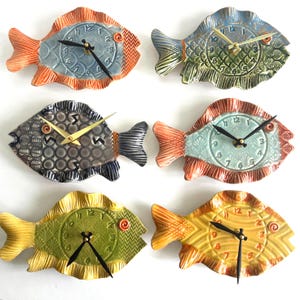 May include: Six ceramic fish-shaped wall clocks arranged in two columns. Each clock features a unique color palette including orange, blue, green, grey, and yellow, with textured scales, a central clock face with numbers, and black or gold hands.