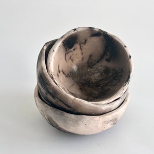 May include: A stack of four handmade ceramic bowls. The bowls are a light beige color with dark gray and black markings. The bowls have a rough, textured appearance and are stacked on top of each other.