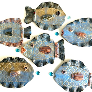 May include: Six ceramic fish sculptures in shades of blue, brown, and gold, arranged on a white surface. Each fish has detailed scales, fins, and eyes. Decorative glass beads are scattered around the fish.