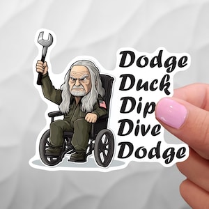 May include: A cartoon sticker of a man in a wheelchair holding a wrench. The man has a long white beard and is wearing a green jacket with an American flag patch. The sticker also includes the words "Dodge Duck Dip Dive Dodge" in black text.