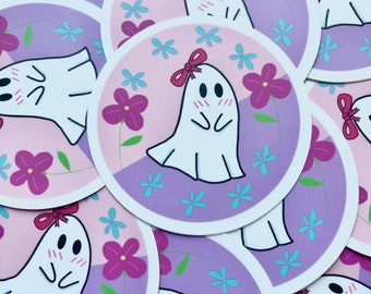 Puede incluir: Un conjunto de pegatinas redondas con un lindo fantasma de dibujos animados con un lazo rosa, mejillas sonrojadas y una expresión tímida. El fantasma está rodeado de flores rosas y moradas sobre un fondo morado claro.