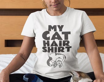 May include: White t-shirt with the text "MY CAT HAIR SHIRT" in black, with a graphic of a cat. The shirt is worn by a person sitting on a bed with a cat. The cat is gray and white.