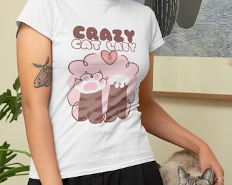 May include: White t-shirt with the text "CRAZY CAT LADY" in brown letters. The shirt features a graphic of cat paws and a heart. The design is in shades of pink and brown. The shirt is being worn by a person.